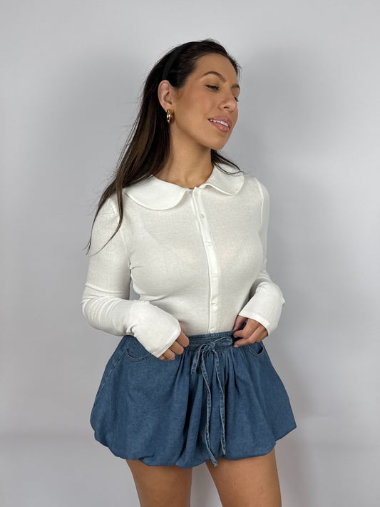 Woman wearing a white pointelle long sleeve top with a Peter Pan collar and blue denim shorts on a plain background