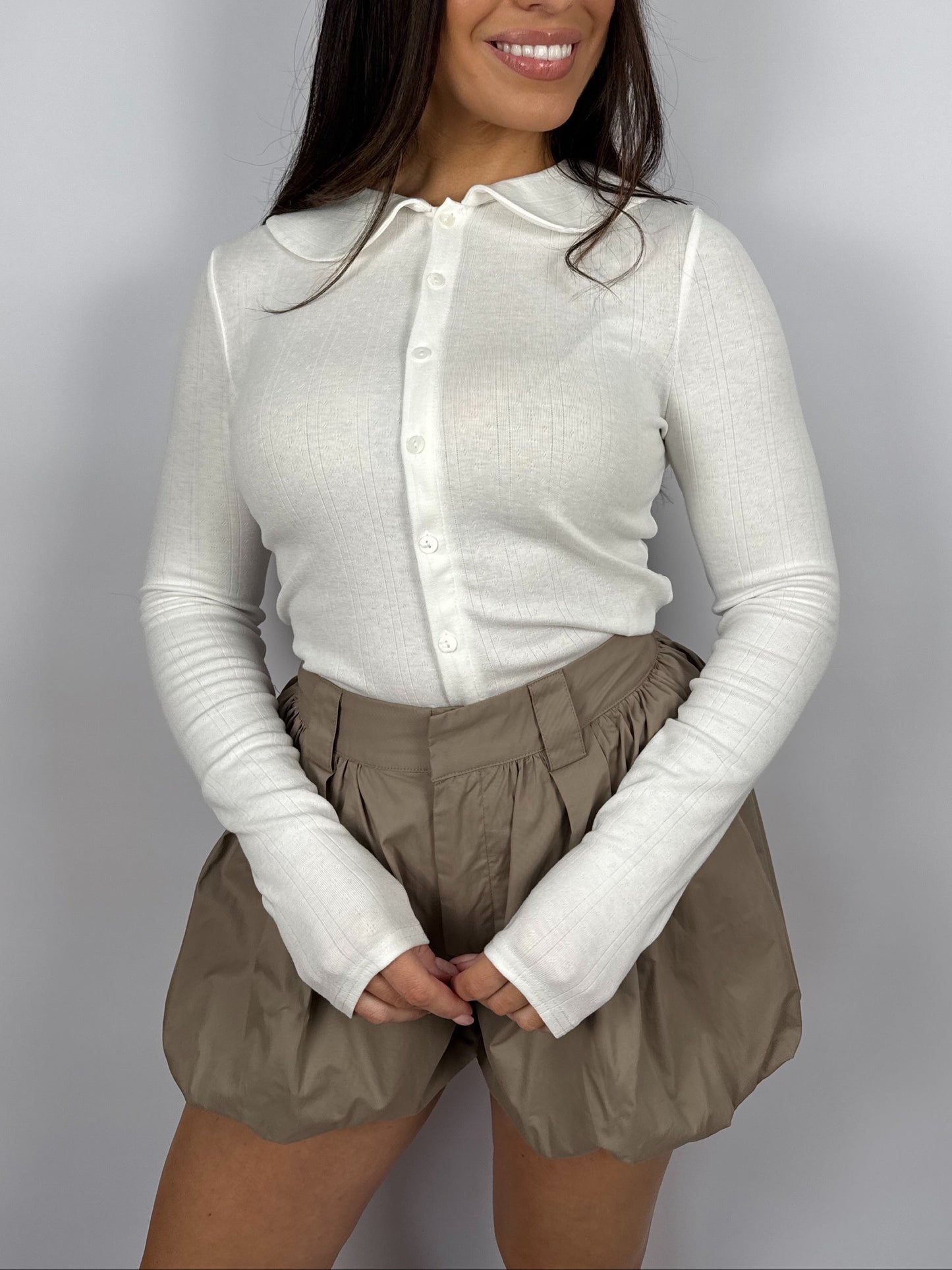 Woman wearing a white pointelle long sleeve top with a Peter Pan collar and beige bubble hem shorts on a plain background