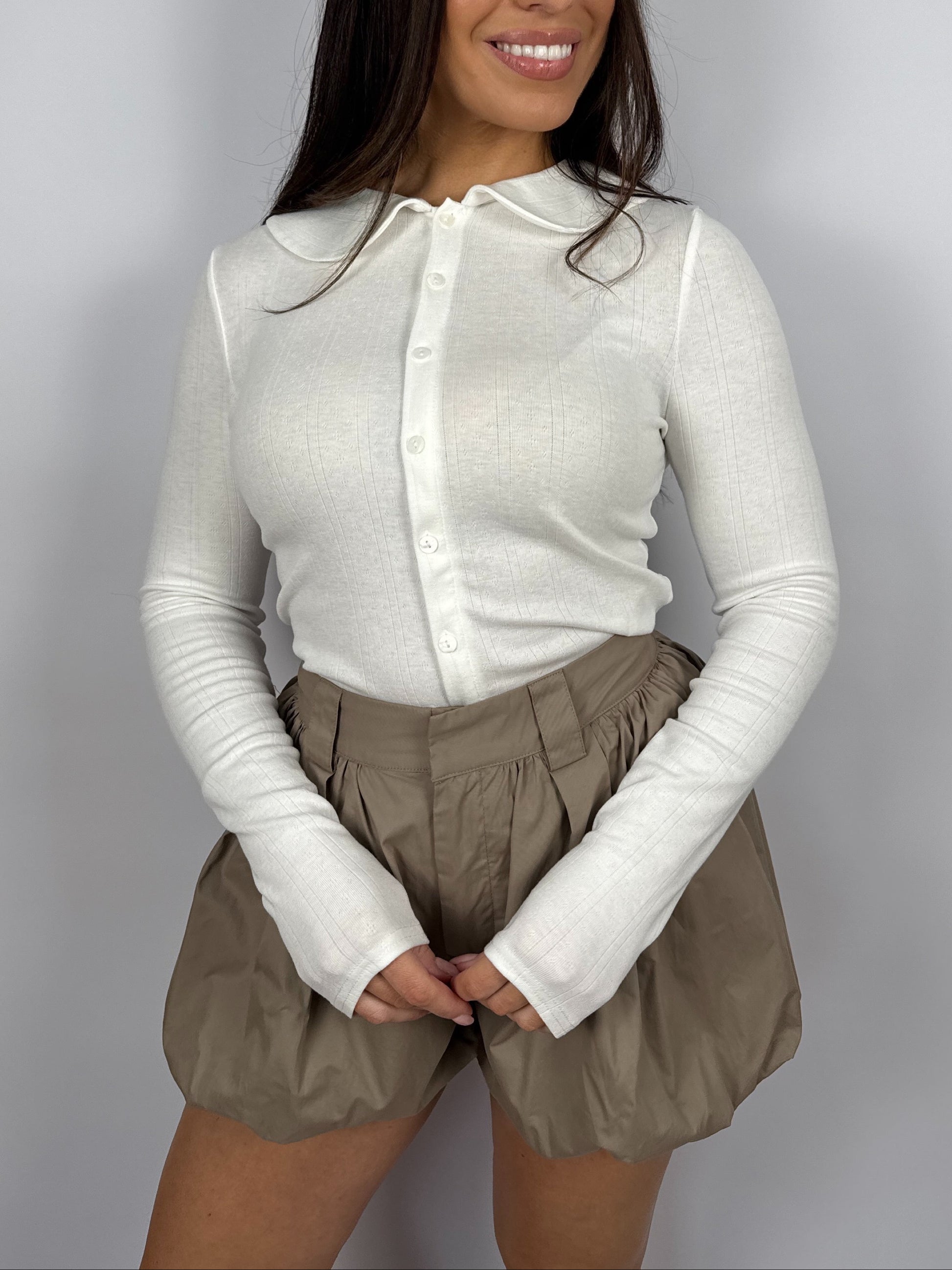 Woman wearing a white pointelle long sleeve top with a Peter Pan collar and beige bubble hem shorts on a plain background