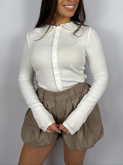 Woman wearing a white pointelle long sleeve top with a Peter Pan collar and beige bubble hem shorts on a plain background