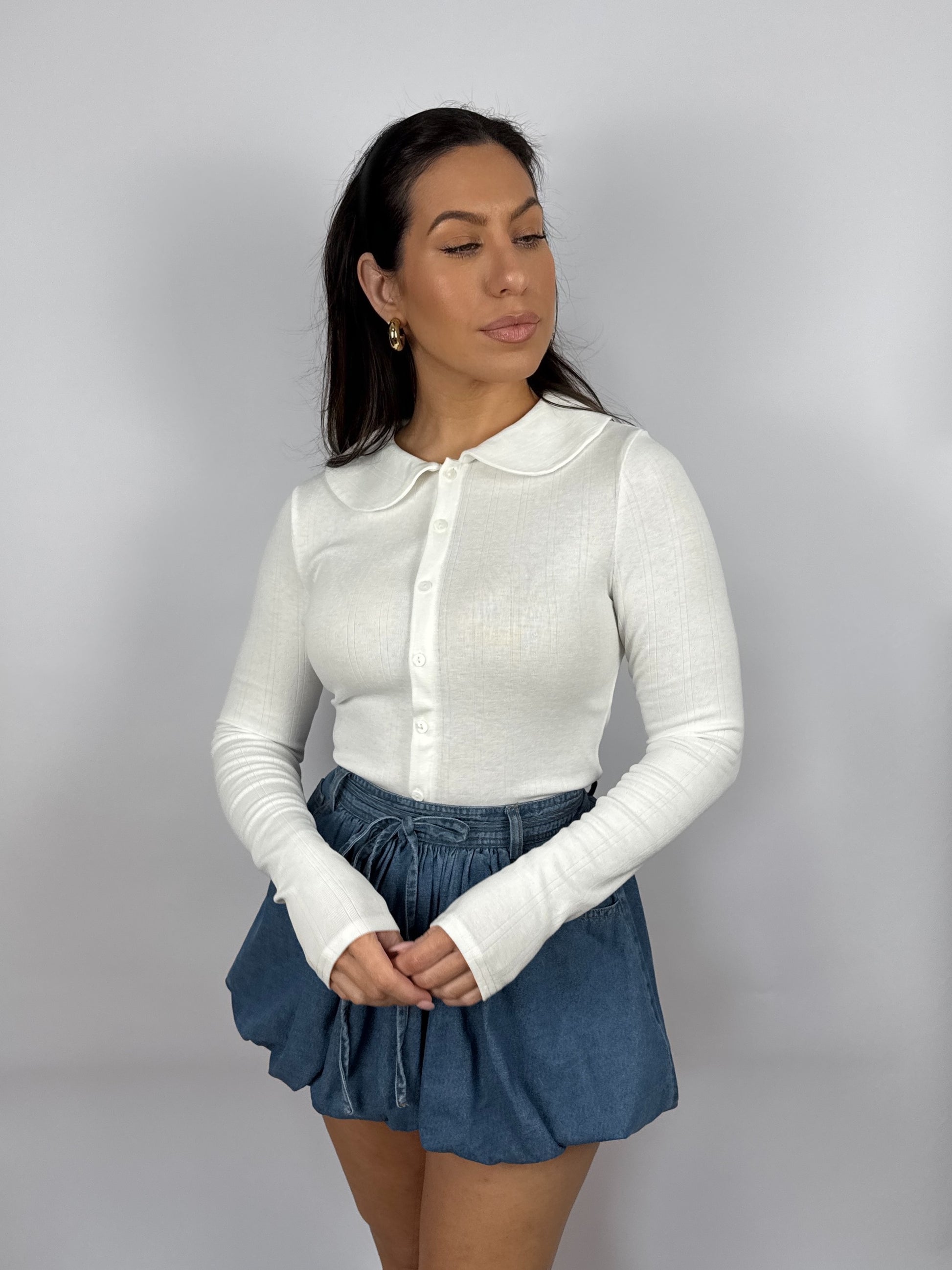 Woman wearing a white pointelle long sleeve top with a Peter Pan collar and blue denim shorts on a plain background