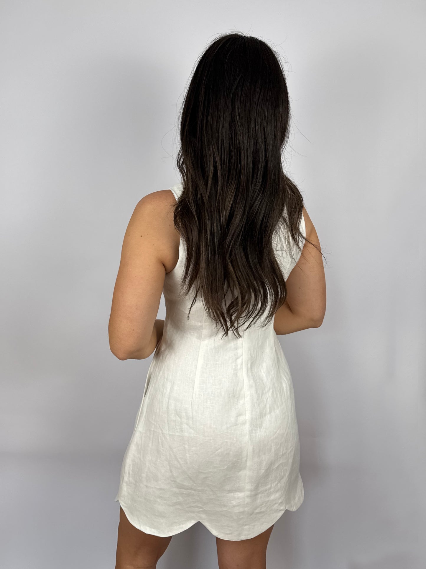 woman wearing a white sleeveless linen dress with scalloped hem against a plain background