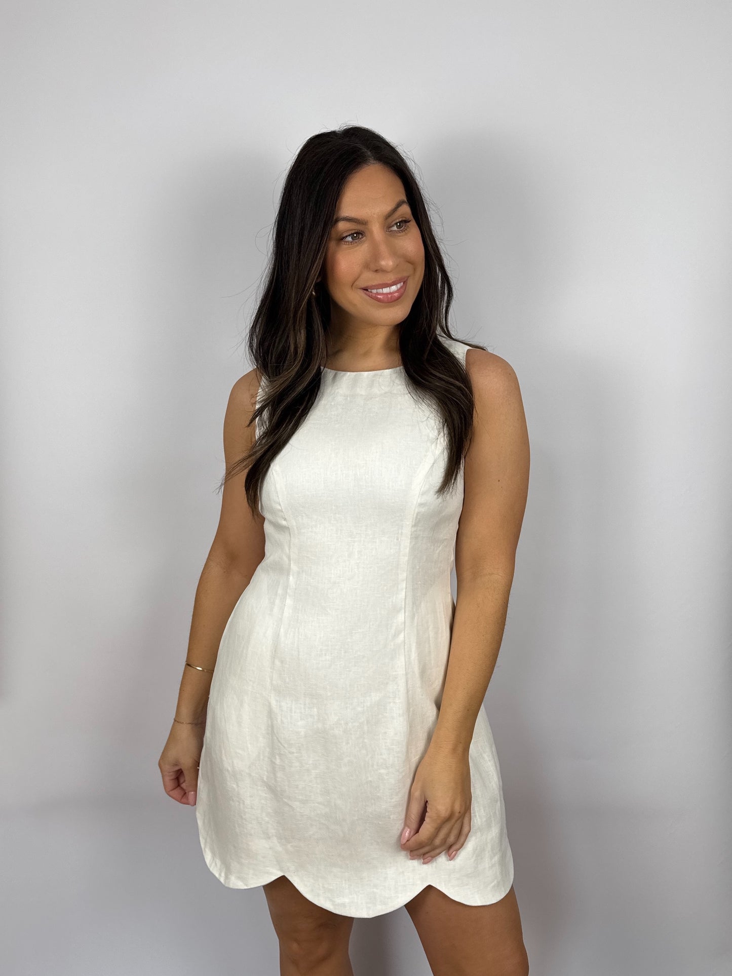 Woman wearing a white linen dress with scalloped hem against a plain background